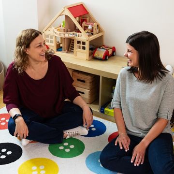 Christina Jungwirth und Magdalena Stark sitzen auf einem bunten Spielteppich und unterhalten sich fröhlich